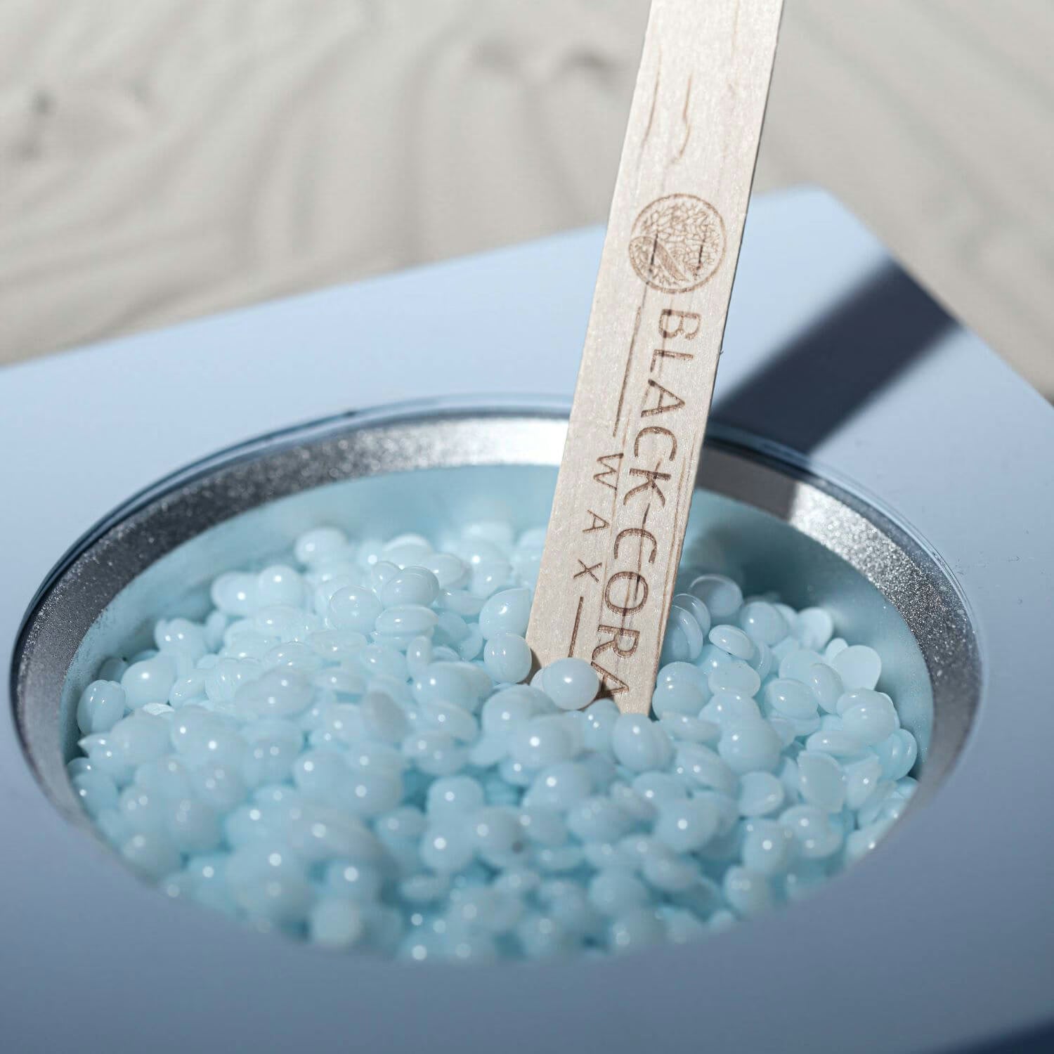 Blue wax beads in a container with a wooden spatula labeled 'Black Core Wax'.